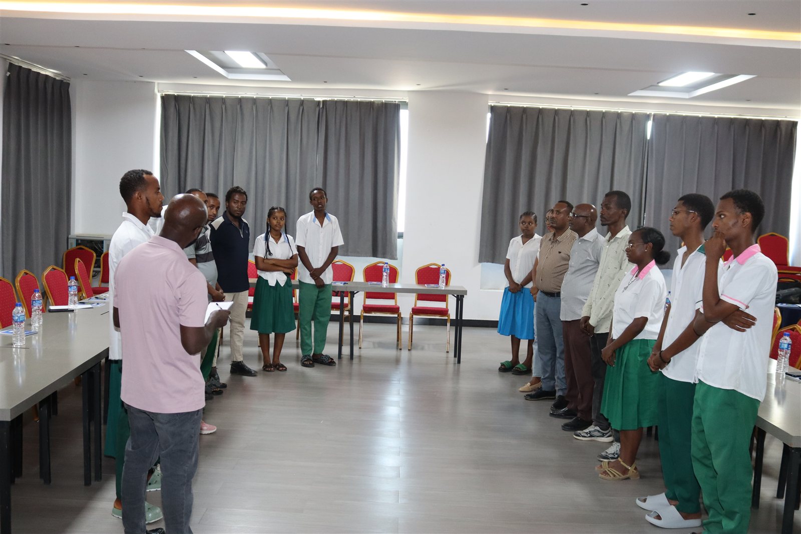 Students in white and green school uniforms and adult facilitators standing in a circle for an opening icebreaker