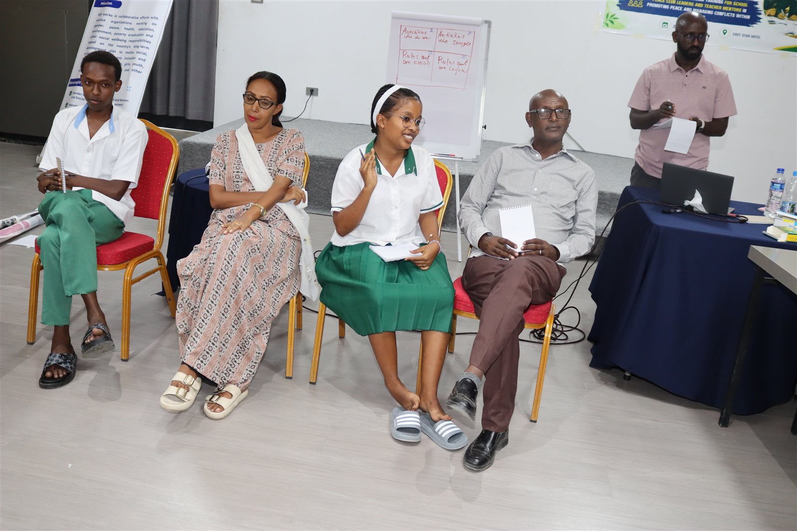 Two students and two adults seated before a flipchart with a four-quadrant self-assessment exercise, part of the event banner visible behind them