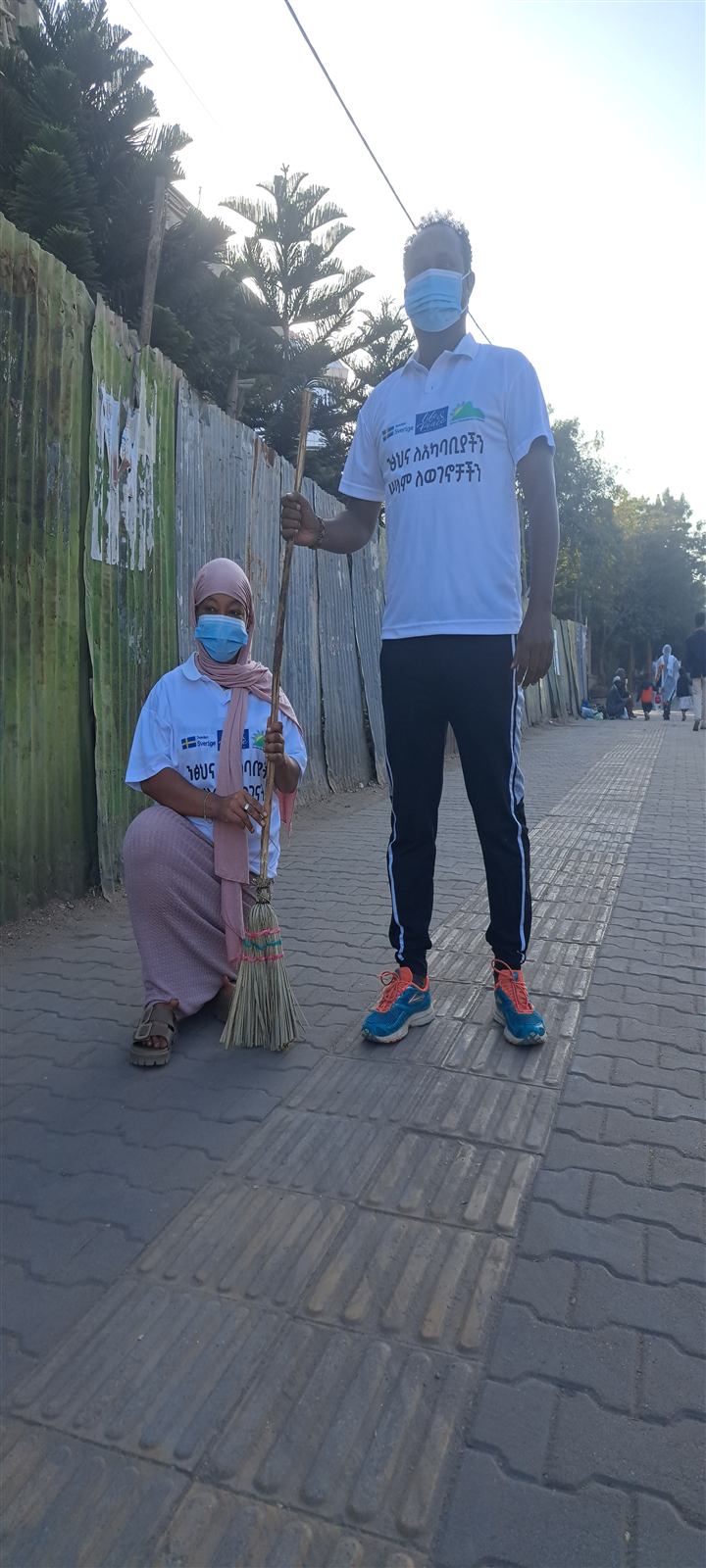 Two women in white campaign T-shirts and face masks sweeping a paved street with a handmade broom