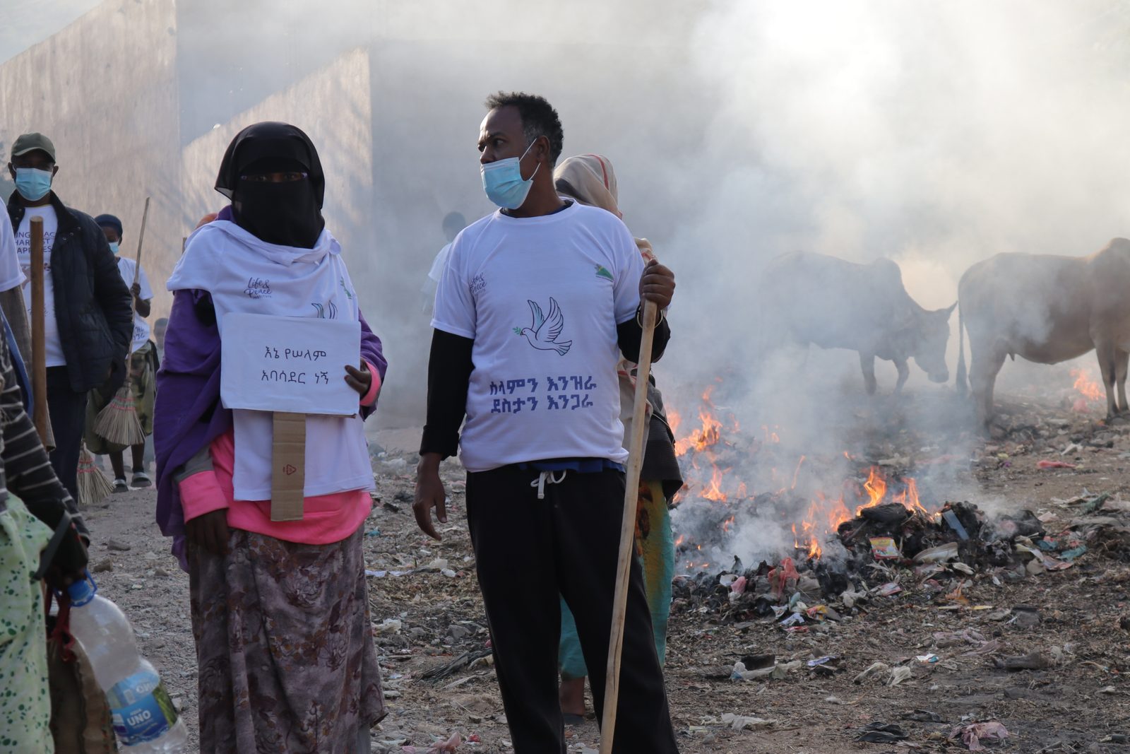 A woman in a black niqab and a man both in white Life and Peace dove-logo T-shirts and surgical masks near a smoldering debris pile