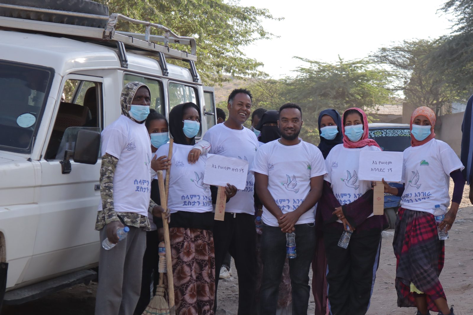 Group beside a vehicle, several masked women holding handwritten Amharic pledge signs and water bottles, smiling